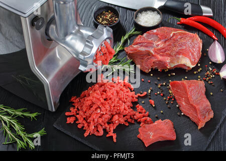 Electric tritacarne è rendere forcemeat di carni fresche bovine carne sul nero ardesia piastra su un tavolo in legno con coltello, Peppe, peperoncino e rosmarino fresco, Foto Stock