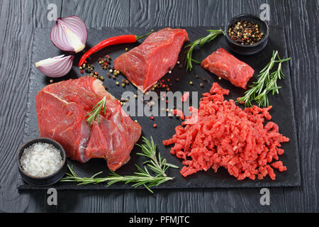 Carni bovine fresche forcemeat e pezzi di filetto di manzo in ardesia nera sulla piastra di tavoli in legno nero con sale, pepe, peperoncino e rosmarino fresco, vista dal Foto Stock