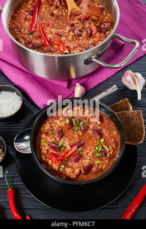 Deliziosi hot chili con carne con tutto il red hot chilis, fagioli, pomodori servito in tazza nera, una pentola e gli ingredienti a sfondo, autentica r Foto Stock