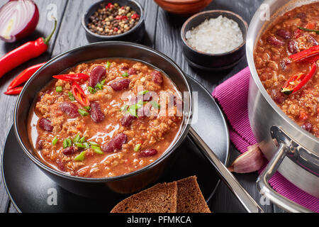 Close-up di deliziosi hot chili con carne con tutto il red hot chilis, fagioli, pomodori servito in tazza nera, una pentola e gli ingredienti a sfondo, Foto Stock