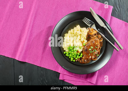 Porzione di hot salato di Salisbury succose bistecche di manzo con cipolla a fungo con sugo di carne servita con purè di patate e piselli verdi su piastre nere con posate, r Foto Stock