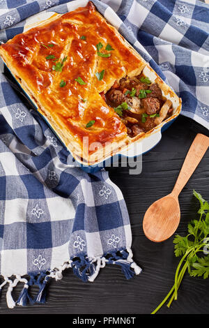 Deliziosa carne chunky e torta di Funghi Conditi con crosta dorata sfoglia di pasta sfoglia in una teglia sul tavolo di legno con carta asciugatutto e cucchiaio di legno Foto Stock