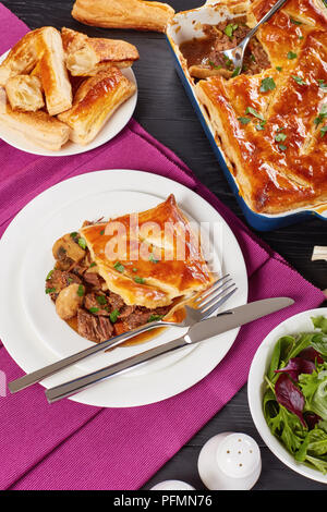 Australian Carni bovine e torta di Funghi Conditi con crosta dorata sfoglia di pasta sfoglia in una teglia e pezzo di torta servita su un piatto con posate e verde Foto Stock