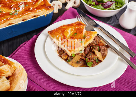 Close-up di manzo australiano e torta di Funghi Conditi con crosta dorata sfoglia di pasta sfoglia in una teglia e pezzo di torta servita su una piastra con cutle Foto Stock