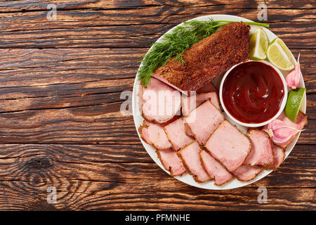 Deliziosi affettati conditi con le spezie con salsa barbecue, fettine di lime, aglio e aneto servita su piastra bianca su tavola in legno rustico, vista Foto Stock