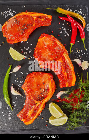 Materie costolette di maiale preparato per cucinare, marinato con spezie e rosso salsa sriracha sul nero ardesia vassoio con peperoncino, sale, fettine di lime e spezie, ver Foto Stock