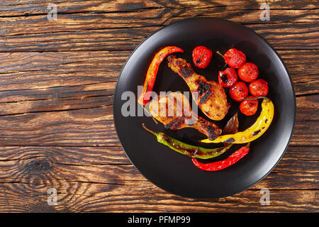 Pollo grigliato con bacchette varicolored il peperoncino e i pomodori ciliegia servito su una piastra nera, vista da sopra, close-up Foto Stock