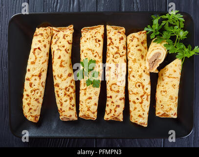 Crepes fritto farcito con pollo tritato e il piatto di formaggi, decorate con prezzemolo fresco lascia su di una piastra nera su tavoli in legno nero, vista da abo Foto Stock