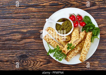 Fritto delizioso Crepes ripiene di carne di pollo servito su una piastra con spinaci, pomodori e salsa pesto, vista da sopra, laici piatta Foto Stock