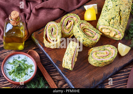 Close-up di deliziosi involtini di zucchine con formaggio fuso e prosciutto bollito tagliato a fettine su un tagliere con saporita salsa di yogurt e gli ingredienti su Foto Stock