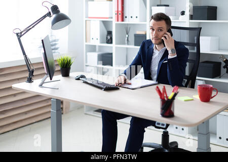 Un giovane uomo è seduto in ufficio al tavolo, parlando al telefono e tenere una matita in mano. Il giovane uomo ha documenti. Foto Stock