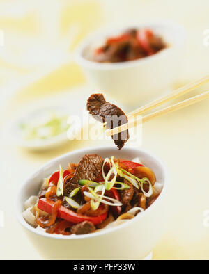 Striscia di stir-fried carni bovine detenute fino con bacchette sopra un piccolo recipiente contenente la carne e le verdure rosolare, vicino. Foto Stock