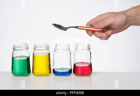 Persona in possesso di cucchiaio sopra quattro barattoli di vetro contenenti verde, giallo, blu e rosso di liquidi Foto Stock