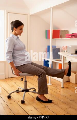 Donna incinta seduta sulla sedia girevole stretching di una gamba Foto Stock