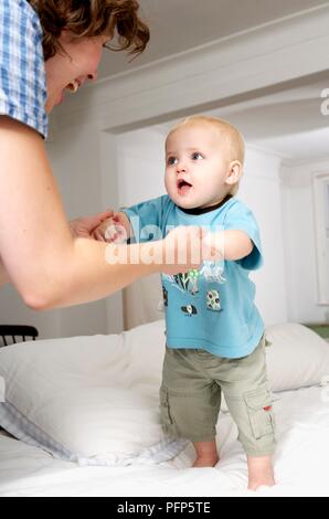 Bambino in piedi sul letto supportato da una donna di mani Foto Stock