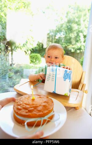 Baby boy in seduta alta apertura dei capelli di un presente, mani tenendo una torta con una sola candela accesa in medio Foto Stock