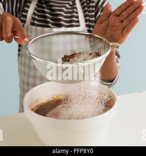 Ragazzo setacciare la farina, il cacao e il lievito in polvere sulla miscela di cioccolato nel recipiente di miscelazione (rendendo brownie al cioccolato), close-up Foto Stock