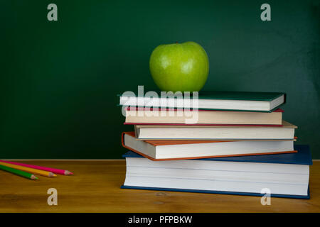 Un insegnante di scuola scrivania con pila di quaderni matite colorate e mela verde. Un verde di Blackboard Foto Stock