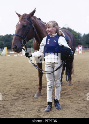 Giovane ragazza nel paddock indossando abito di equitazione in piedi accanto pony, tenendo redini e hat Foto Stock