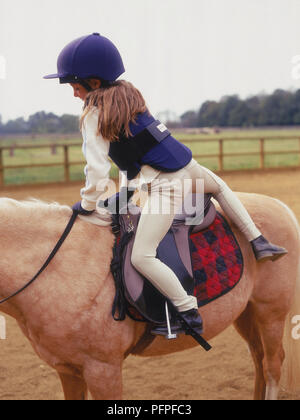 Ragazza giovane indossando abito di equitazione, montaggio palomino pony nel paddock Foto Stock