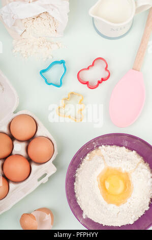 La cottura Ingredienti e utensili, inclusi cartone di uova, le uova e la farina nel recipiente di miscelazione, brocca del latte, farina fuoriuscita dal sacco, pasticceria taglierine e miscelazione di cucchiaio, vista aerea Foto Stock