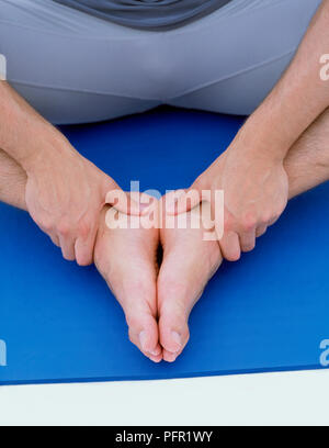 Uomo seduto sul tappeto blu, suole di toccare i piedi e le mani sulle caviglie, close-up (caviglia stretch) Foto Stock