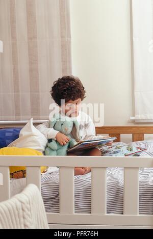 Tre anni di vecchio ragazzo seduto sul letto azienda giocattolo morbido e guardando il libro Foto Stock