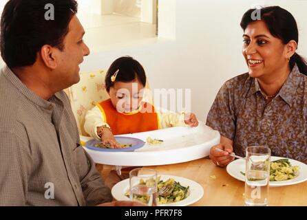 L uomo e la donna seduta a tavola chiacchierando a cena, bimbo seduto con loro nel seggiolone con in mano una ciotola di cibo Foto Stock