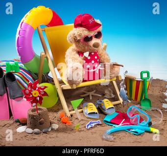 Orsacchiotto di peluche seduto sulla sedia a sdraio sulla spiaggia indossando il costume da bagno, occhiali da sole e cappello e tenendo il gelato Foto Stock