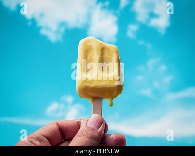 Mano che tiene il giallo congelati ghiaccioli di ghiaccio in fusione Pop su estate cielo blu sullo sfondo Foto Stock
