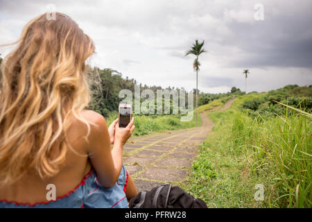 Bella giovane donna in vestito blu sedile in pietra percorso in salita in Asia e scattare foto sul suo smartphone. Ragazza di viaggio e di esplorare il mondo. Long lonely ro Foto Stock