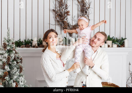 Amorevole famiglia un Buon Natale e Felice Anno Nuovo. Allegro piuttosto persone. Mamma e papà abbracciando piccola figlia . I genitori e il bambino bambino divertirsi nei pressi di albero di Natale e caminetto bianco all'interno. Foto Stock