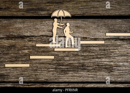 Due piccole silhouette di persone a camminare su scala come uno detiene un ombrello sopra gli altri come si cammina su uno sfondo di legno. Foto Stock
