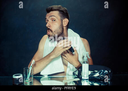 Il giovane uomo in camera da letto seduto di fronte allo specchio di graffiare la sua barba a casa. Le emozioni umane concept Foto Stock