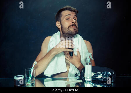 Il giovane uomo in camera da letto seduto di fronte allo specchio di graffiare la sua barba a casa. Le emozioni umane concept Foto Stock