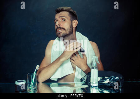 Il giovane uomo in camera da letto seduto di fronte allo specchio di graffiare la sua barba a casa. Le emozioni umane concept Foto Stock