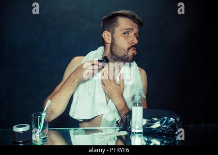 Il giovane uomo in camera da letto seduto di fronte allo specchio di graffiare la sua barba a casa. Le emozioni umane concept Foto Stock