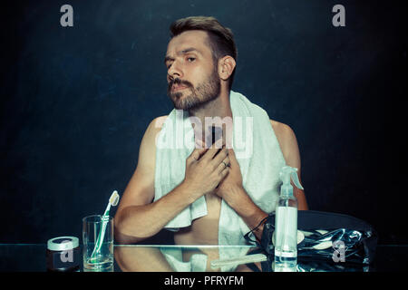 Il giovane uomo in camera da letto seduto di fronte allo specchio di graffiare la sua barba a casa. Le emozioni umane concept Foto Stock