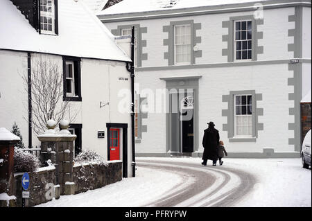 Un uomo che indossa un soprabito e un grande cappello cammina attraverso una strada innevata a Kirkcudbright, Dumfries e Galloway, Scozia, Regno Unito, con un bambino piccolo Foto Stock