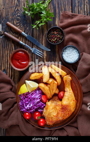 Patate fritte cunei ,Pollo gamba e coleslaw insalata servita su una piastra di argilla rustico con coltello e forchetta al buio su un tavolo di legno, vista verticale da abov Foto Stock