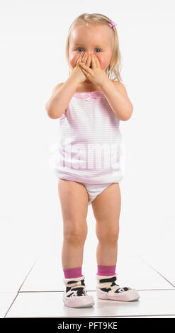 Ragazza indossando giubbotto, mutande e pantofole, che copre il volto con le mani, 20 mesi Foto Stock