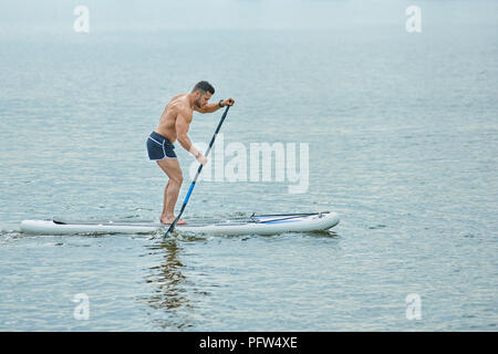 Giovane uomo con corpo sportivo in piedi sul bordo del pannello SUP, il roaming con una pozzanghera. Sportivo a mantenere uno stile di vita sano, nuoto nel lago della città. indossare camicie nere, avente forti muscoli. Foto Stock