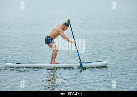 Montare l'uomo avente la formazione con sup board, il roaming con lunghi pozza nel lago della città. Modello maschile indossare pantaloncini sport, permanente sulla grande lungo bordo, nuotare sulla superficie dell'acqua. Coltivare uno stile di vita sano. Foto Stock