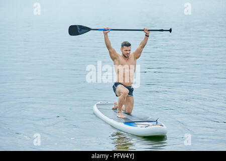 Vista frontale di uno sportivo mantenendo lungo remo sopra la testa, nuoto sul sup pensione in città sul lago. Modello maschile avente fit con forti muscoli, mantenendo uno stile di vita sano. Formazione durante la fase estiva soleggiata giornata. Foto Stock