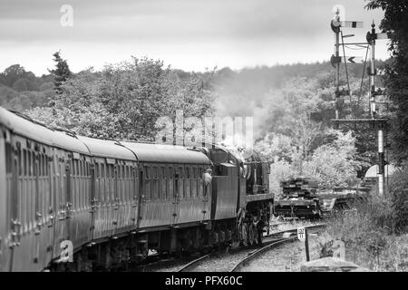 Vista laterale in bianco e nero del treno a vapore d'epoca britannico che tira le carrozze che viaggiano in pista oltre i segnali ferroviari, linea storica della Severn Valley Railway. Foto Stock