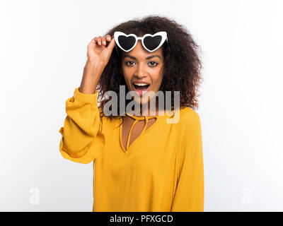 Bella americano africano donna con capelli afro in giallo e indossare occhiali da sole sorride alla telecamera oltre il muro bianco sullo sfondo. Carino razza mista ragazza por Foto Stock