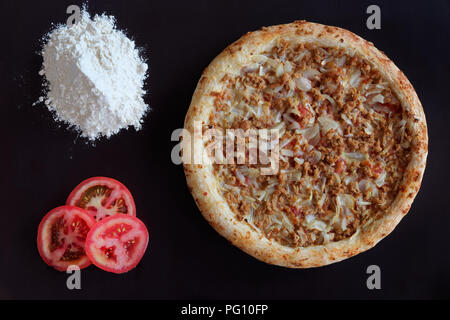 Tonno al forno pizza, mucchio di farina e fette di pomodoro su sfondo nero Foto Stock