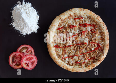 Tonno al forno pizza, mucchio di farina e fette di pomodoro su sfondo nero Foto Stock