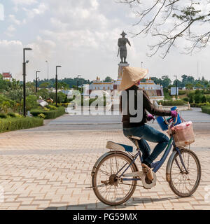 Un laotiano donna corse in bicicletta in Chao Anouvong park con la grande statua del re Chao Anouvong in background in Vientiane, Laos Foto Stock