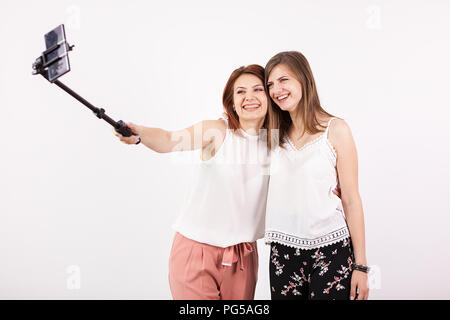 Due bella giovane donna prendendo un selfie con un bastone selfie e produrre diverse divertenti e sciocco volti mentre facendo. Studio foto su un muro bianco Foto Stock